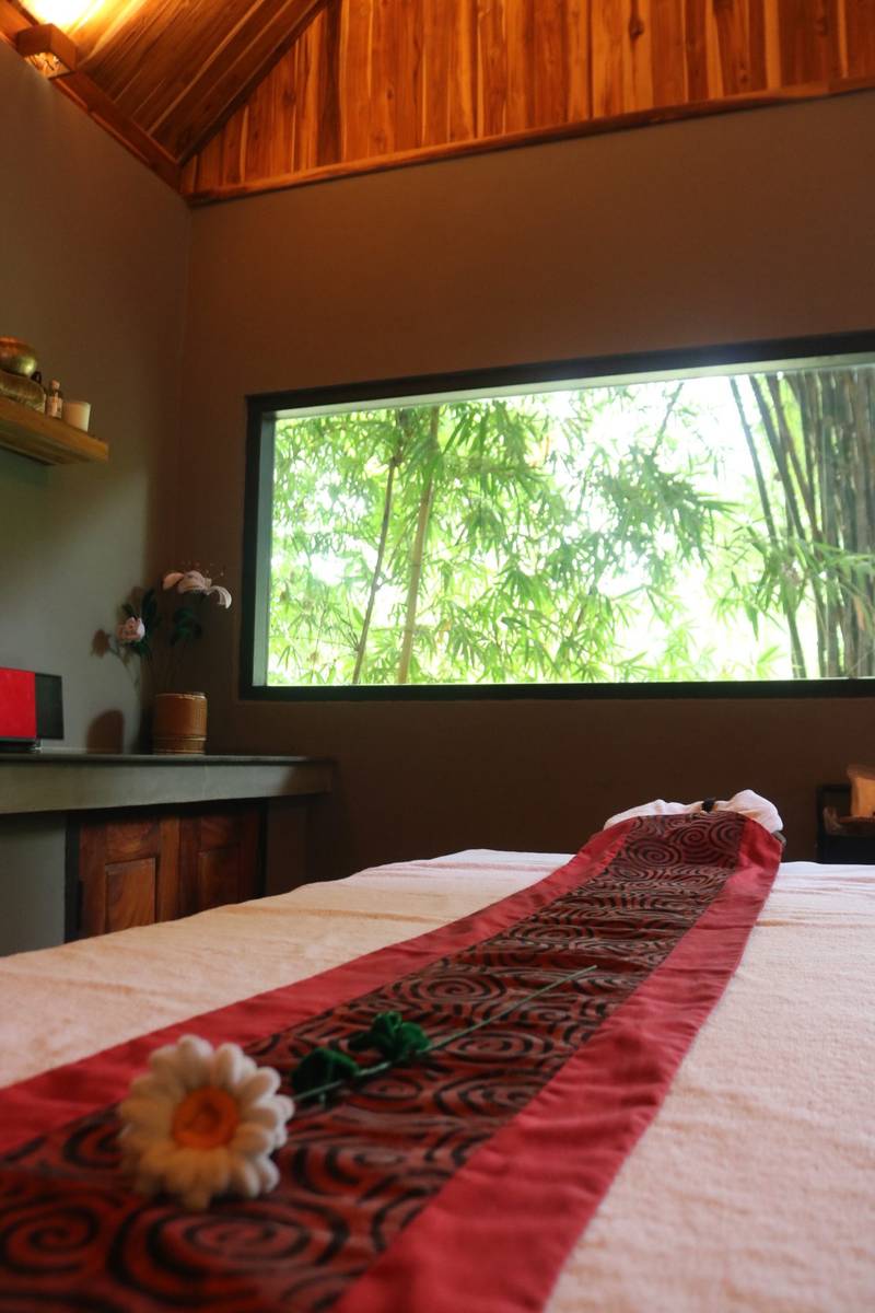 Spa treatment room at The Namkhan in Luang Prabang featuring massage bed with red silk runner and bamboo garden views through picture window