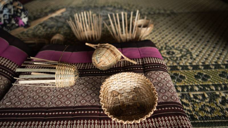 Handwoven bamboo baskets rest on traditional Lao textiles with intricate geometric patterns in burgundy, black, and natural tones — The Namkhan Luang Prabang