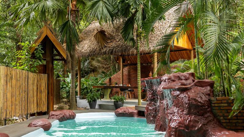 The Namkhan pool surrounded by lush tropical palms and natural rock waterfall features in Luang Prabang, with traditional thatched pavilions