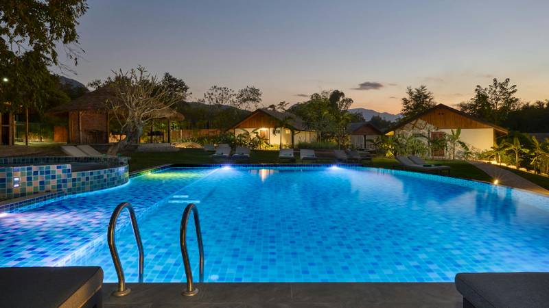 Illuminated swimming pool at dusk with glowing resort bungalows and mountains beyond.