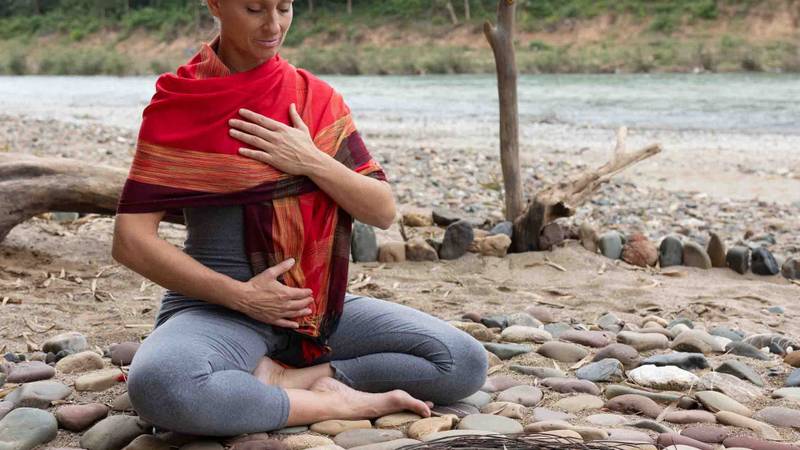 Guest practicing meditation on the riverbank with traditional Lao textiles, surrounded by smooth stones and driftwood along the water's edge