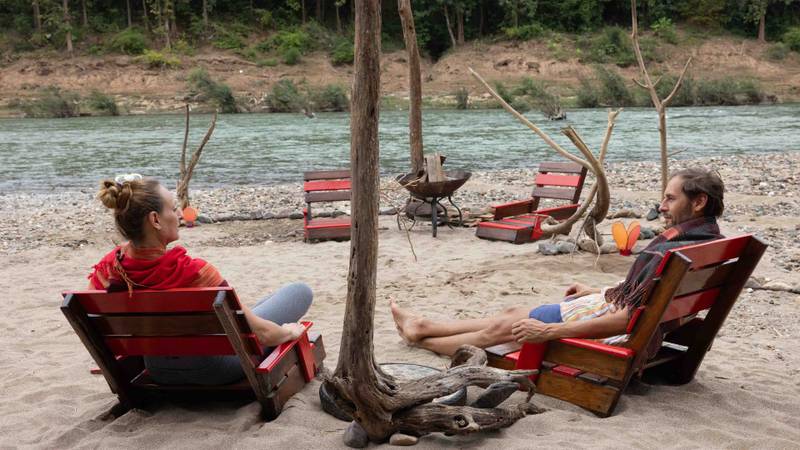 Two guests relax in red wooden chairs around a fire pit on a sandy riverside beach with the Nam Khan River and jungle backdrop