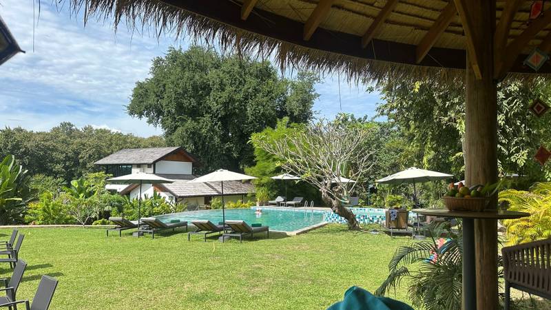 View of The Namkhan resort pool surrounded by manicured lawns and tropical gardens, framed by traditional thatched sala in Luang Prabang, Laos