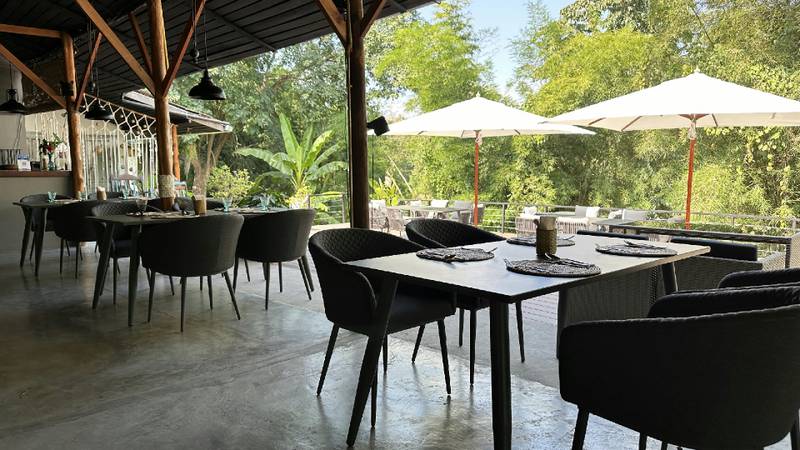 Empty dining tables with black chairs on an open-air restaurant terrace surrounded by greenery.