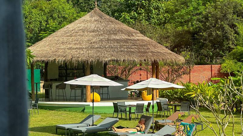Hotel pool with mosaic tiles, sun loungers on manicured lawn, thatched-roof pavilion bar, and mountain backdrop under blue skies