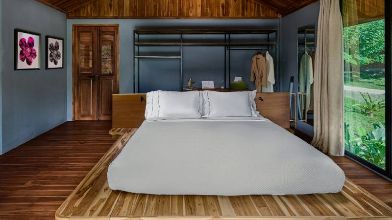 A neatly made white bed in a teak-panelled suite with open wardrobe behind.