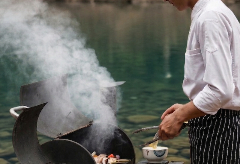 Chef in traditional white uniform grilling fresh ingredients on smoky outdoor barbecue beside tranquil river at Namkhan resort