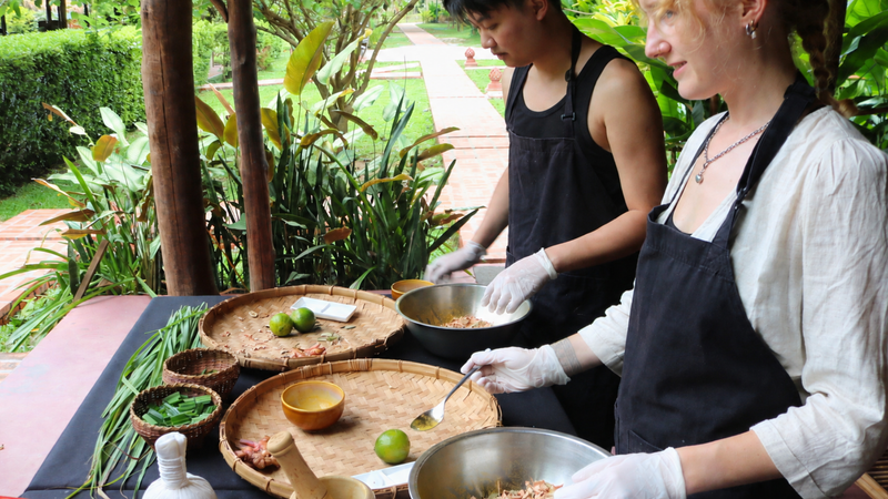 Two guests in aprons prepare traditional Lao dishes with fresh herbs and mortar-pestle at The Namkhan cooking class in Luang Prabang, Laos