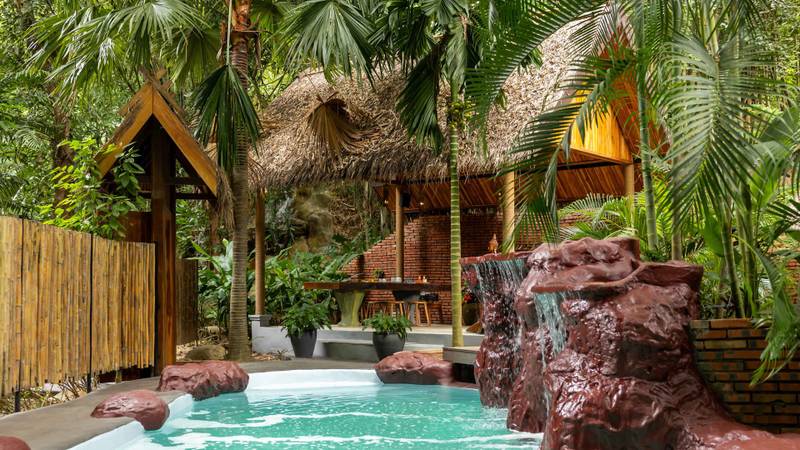 Private resort pool surrounded by lush palm trees and tropical foliage, featuring natural rock waterfall and traditional thatched pavilion