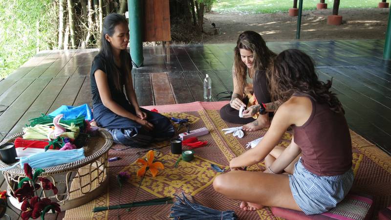 Three guests sitting on woven mat learning traditional Lao craft-making with local instructor in open-air pavilion at Namkhan ecolodge