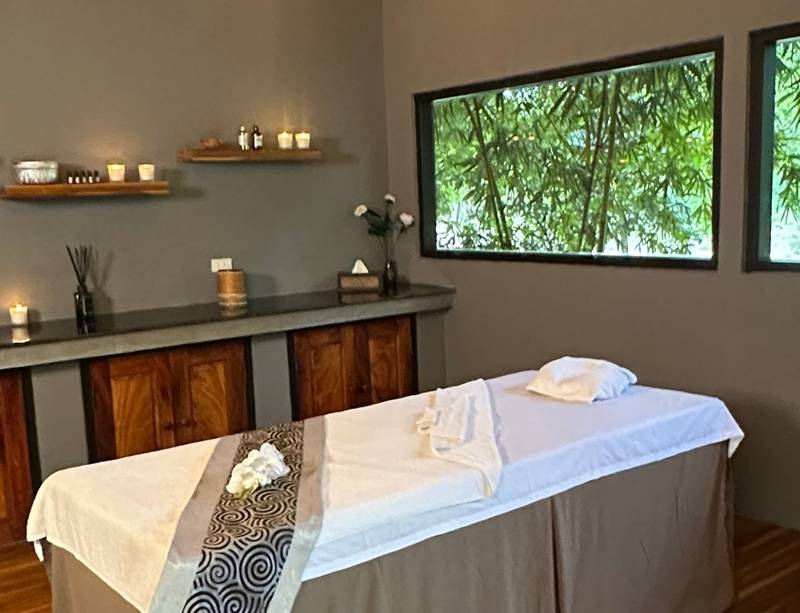 Tranquil spa treatment room at The Namkhan in Luang Prabang featuring vaulted bamboo ceiling, massage bed, and jungle views through picture windows.