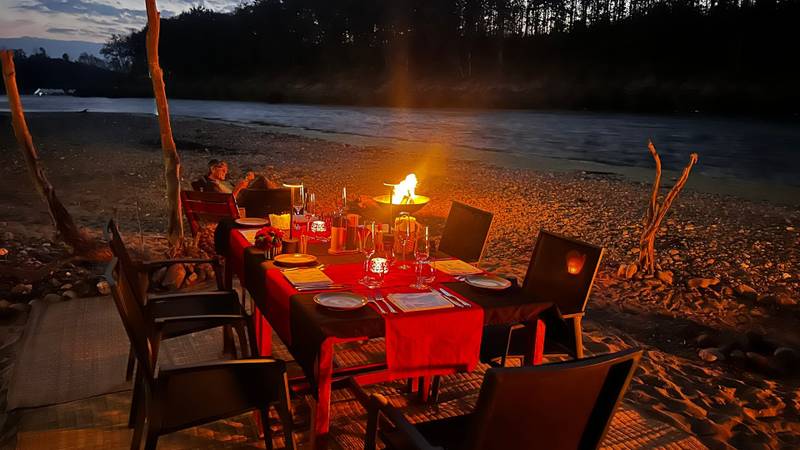 Intimate riverside dining setup with firepit at The Namkhan resort in Luang Prabang, featuring candlelit table on pebbled riverbank at dusk