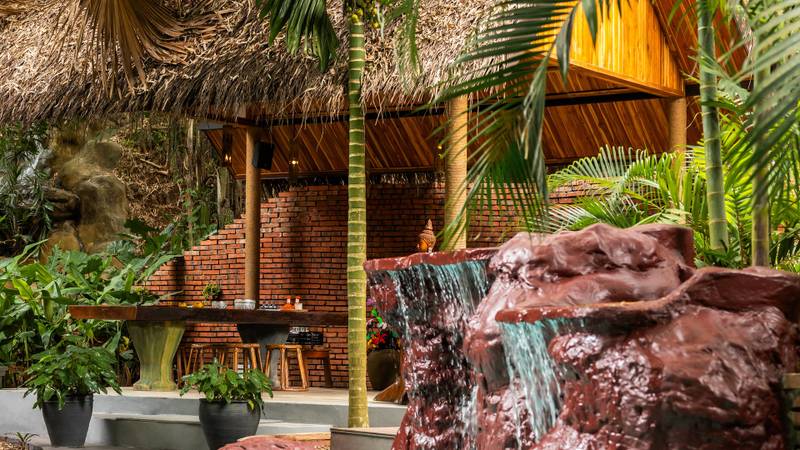 Turquoise pool with cascading waterfall over terracotta rocks, framed by palm fronds and a thatched-roof bar with brick counter