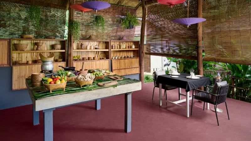 Open-air cooking space with produce-laden table, teak shelving, and a set dining table.