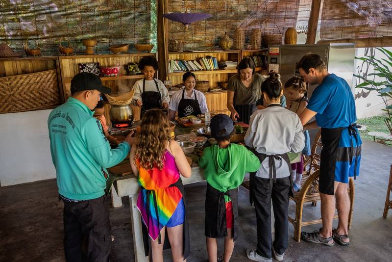 Guests of all ages gather around a cooking station in a traditional bamboo kitchen, learning to prepare authentic Laotian dishes together