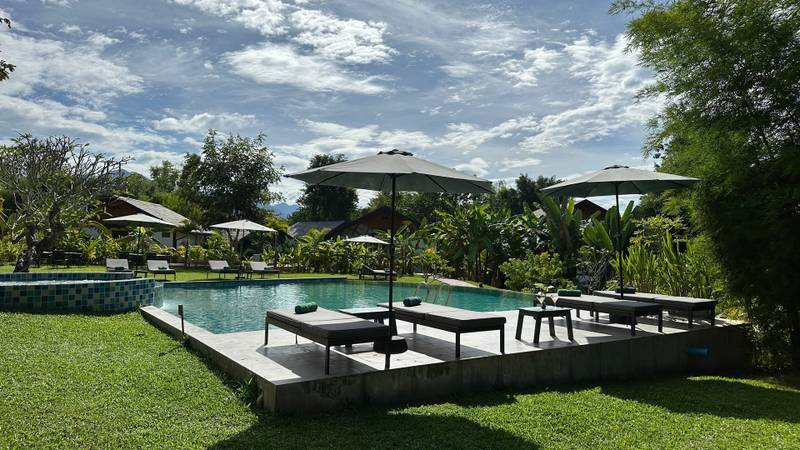 Sunlit pool with dark wooden loungers and white umbrellas surrounded by tropical gardens at The Namkhan resort in Luang Prabang, Laos