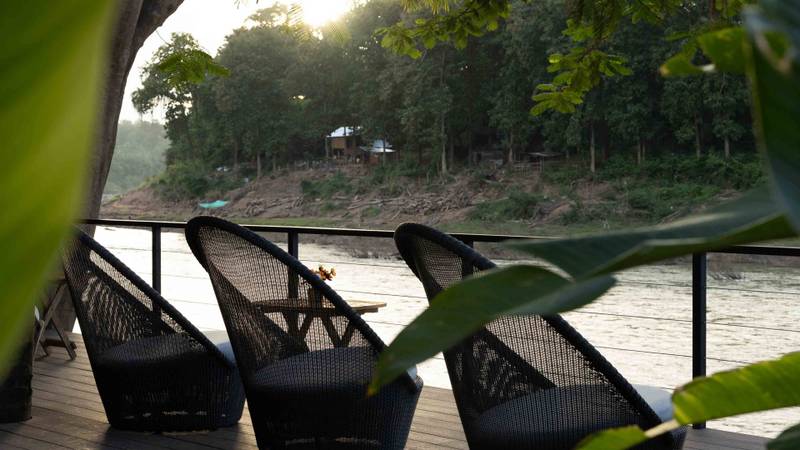 Black woven lounge chairs arranged on wooden deck overlooking Nam Khan River framed by overhanging tree branches at golden hour