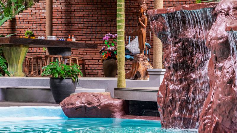Natural rock waterfall cascading into turquoise pool with brick pavilion, wooden Buddha statue, and tropical plants at Namkhan resort