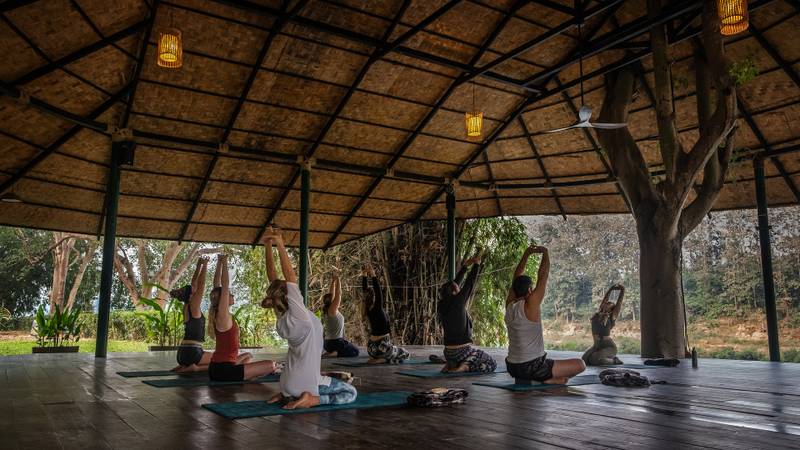 Group yoga class in traditional thatched pavilion with natural light, wooden deck, and views of surrounding gardens at Namkhan resort