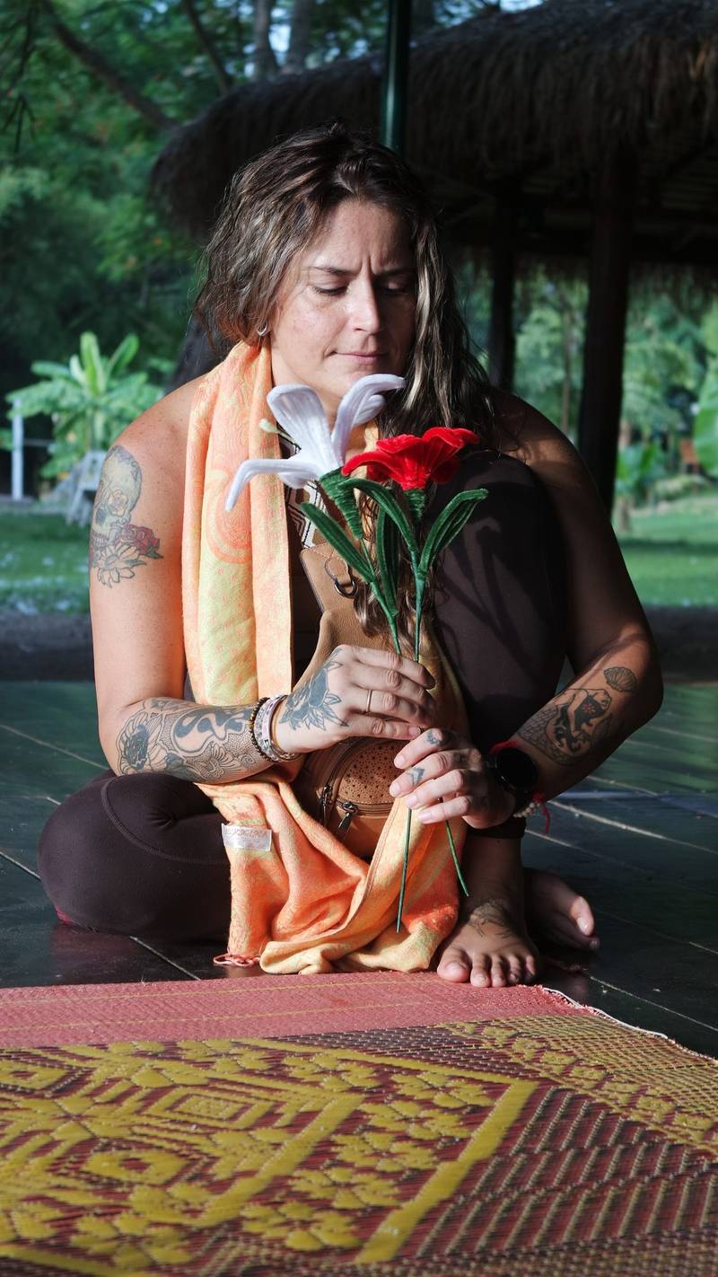 Guest in orange robe sits cross-legged holding lotus flowers during mindfulness ritual at Nam Khan's open-air meditation pavilion