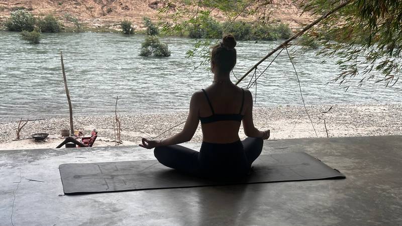 Guest practicing yoga meditation in lotus pose on concrete platform overlooking the Nam Khan River, framed by overhanging willow branches