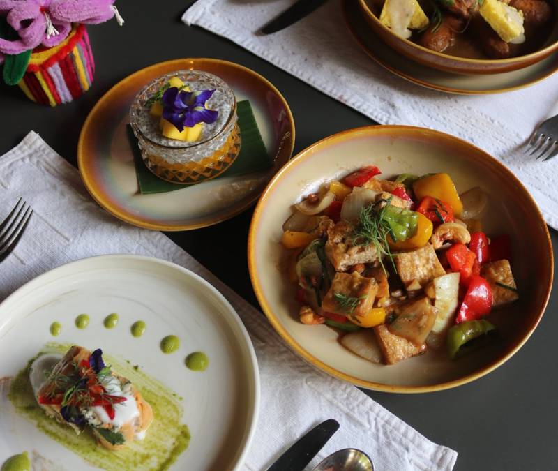 Overhead view of colorful Lao tasting menu dishes with fresh vegetables, herbs, and traditional pottery on white linen table setting — The Namkhan Luang Prabang