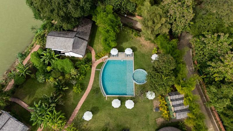 Aerial view of The Namkhan resort pool surrounded by lush tropical gardens along the riverside in Luang Prabang, Laos, with white umbrellas and villa.