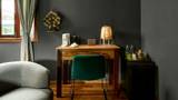 A teak writing desk with rattan lamp and decorative wall piece in a dark-walled room.