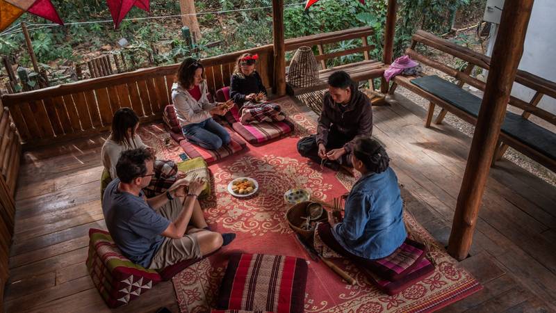 Guests gather on wooden deck for traditional Lao weaving workshop, seated on tribal cushions surrounded by jungle, guided by local artisans — The Namkhan Luang Prabang
